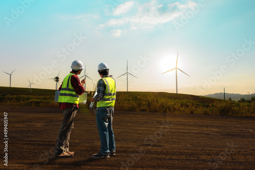 Two maintenance engineer working on wind turbine farm in sunset, discussed plan for the maintenance of wind turbines while holding blueprint of layout plan.