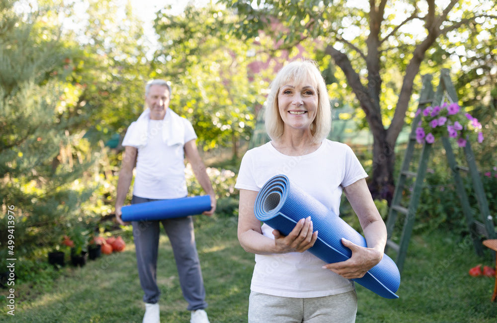 Obraz premium Portrait of active senior woman posing with husband in garden, holding yoga mats, exercising together outdoors