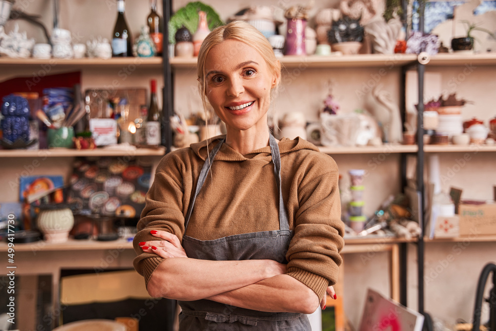 Artist woman wearing apron standing arms crossed at her studio and smiling