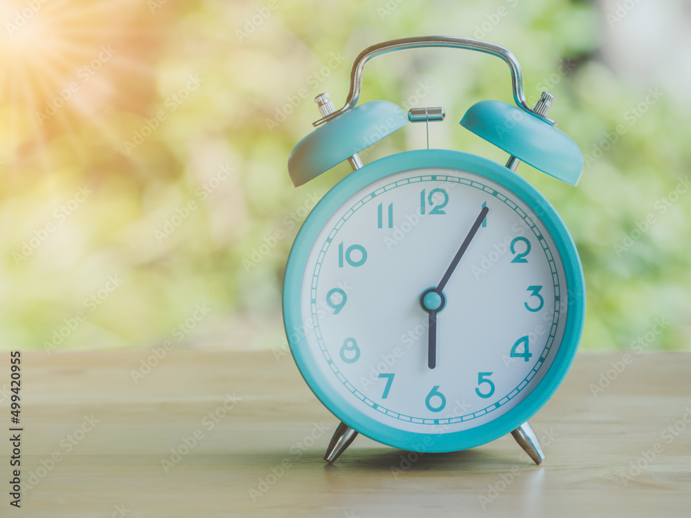 Retro alarm clock 6 o'clock AM or PM with morning sun with flare light blur green background on wooden table. Retro alarm clock 6 o'clock AM or PM with morning sun with flare light blur green background on wooden table.