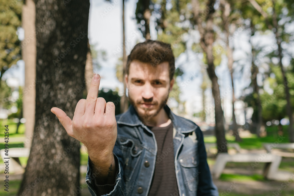 Young man wearing blue jeans jacket over green city park background. Showing middle finger, impolite and rude fuck off expression. Young man wearing blue jeans jacket over green city park background. Showing middle finger, impolite and rude fuck off expression.