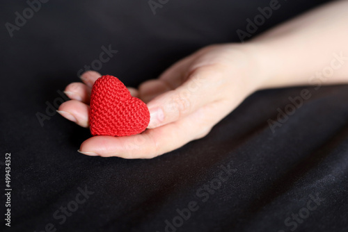 Red knitted heart in female palm of hand on a bed with black bedding. Concept of love, health care, motherhood, blood donation