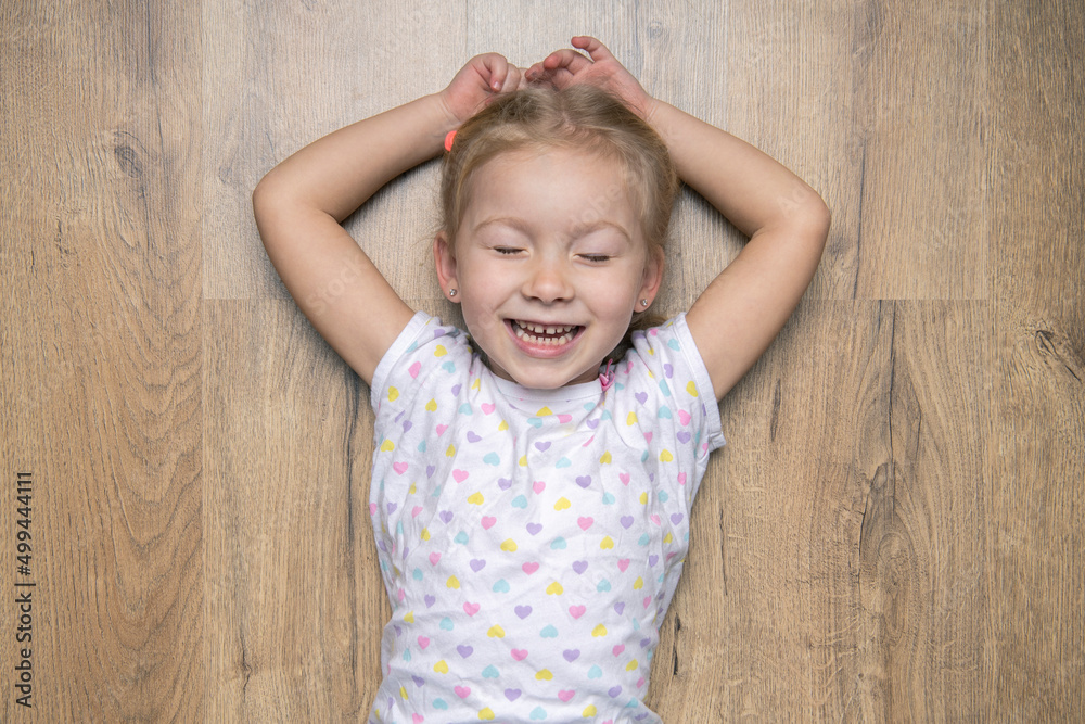 Fototapeta premium Smiling child girl with closed eyes lying on a wooden floor. Top view, flat lay.
