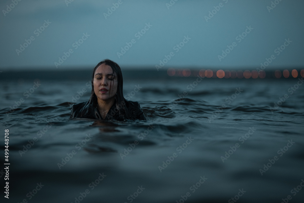beautiful brunette woman stands in a coat in the sea with her eyes closed at night
