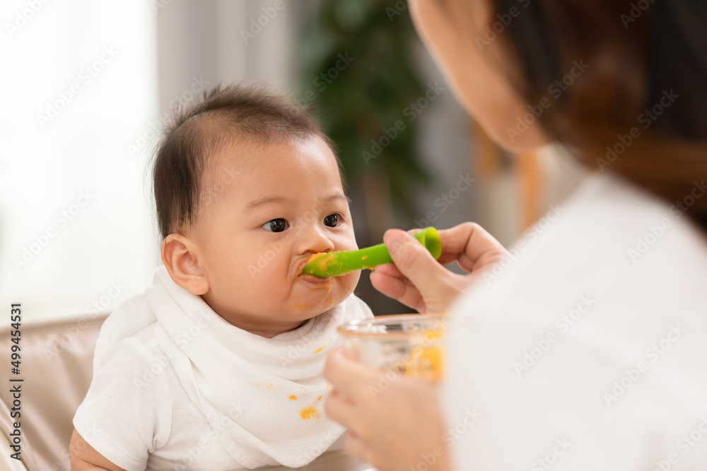 Side view mother feeding her asian baby daughter with pumpkin mashed or vegetable mash on rubber spoon.Mom trying to feed little baby boy at home enjoy and spending time together.Baby feeding Concept Side view mother feeding her asian baby daughter with pumpkin mashed or vegetable mash on rubber spoon.Mom trying to feed little baby boy at home enjoy and spending time together.Baby feeding Concept