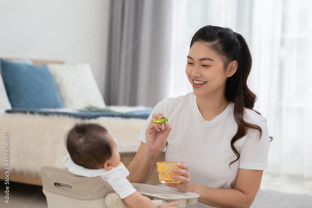 Mother feeding her asian baby daughter with pumpkin mashed or vegetable mash for vitamin on rubber spoon.Mom trying feed little baby boy at home enjoy and spending time together.Baby feeding Concept