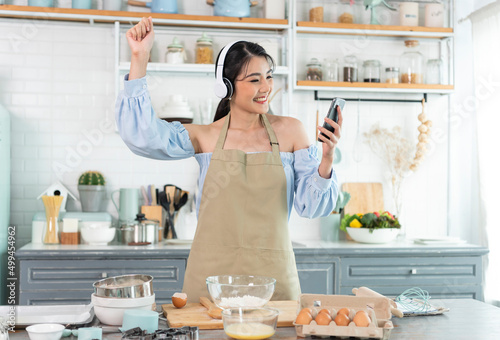 happy cheerful young asian woman raise smartphone fist good mood wear headphones listen to music in kitchen at home.