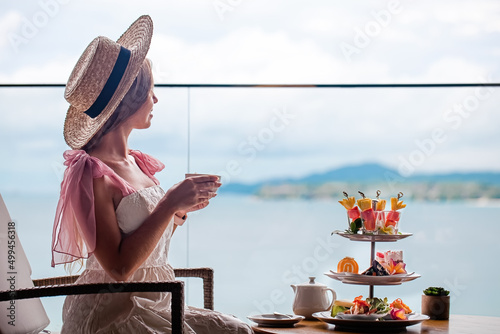 Cheereful young woman in white dress and hat sitting on chair at modern cafe with sea view, drinking hot tea and eating tasty desserts from stand with sweet cakes, enjoying and smiling. Slow motion. photo