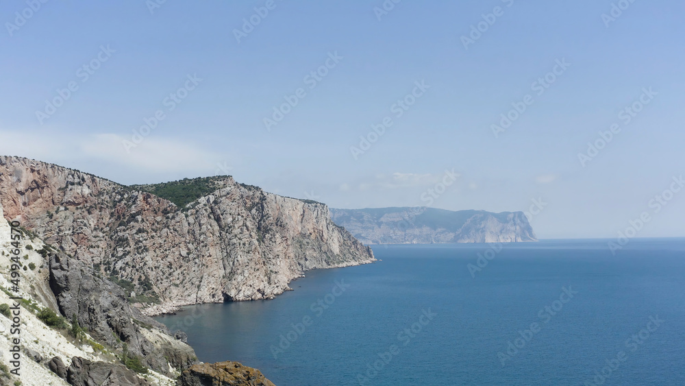 Aerial view of rocky coast and blue sea. Action. Flying above steep slopes of a mountain range, cliffs above calm water surface, summer nature landscape.
