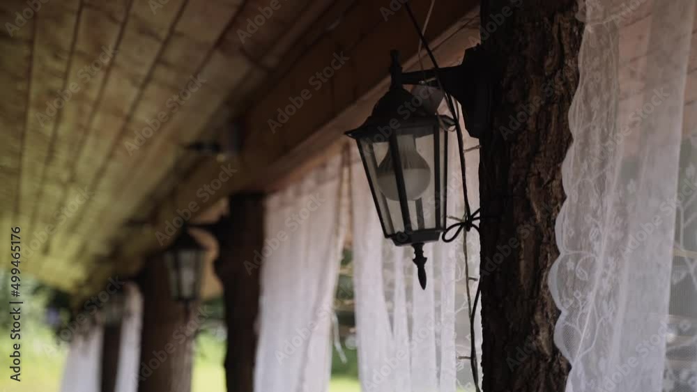 Black glass retro lantern with LED lamp on wooden pole in the form of natural tree trunk with bark on open terrace with curtains, close-up. Illumination of village house in the countryside.