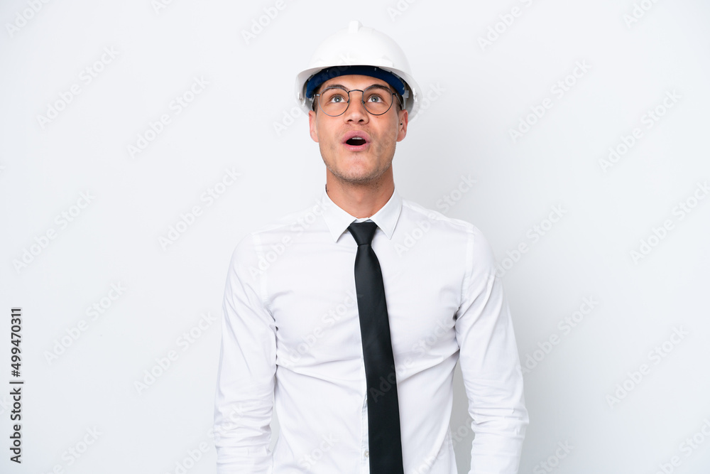 Young architect caucasian man with helmet and holding blueprints isolated on white background looking up and with surprised expression Young architect caucasian man with helmet and holding blueprints isolated on white background looking up and with surprised expression