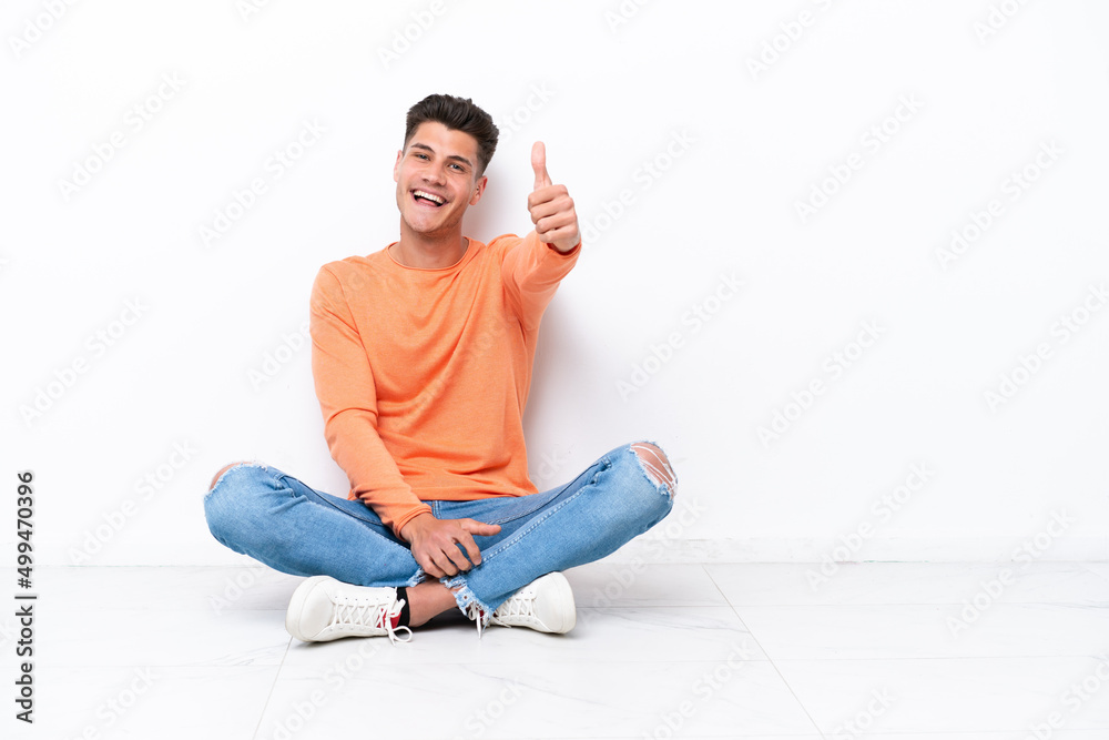 Young man sitting on the floor isolated on white background with thumbs up because something good has happened