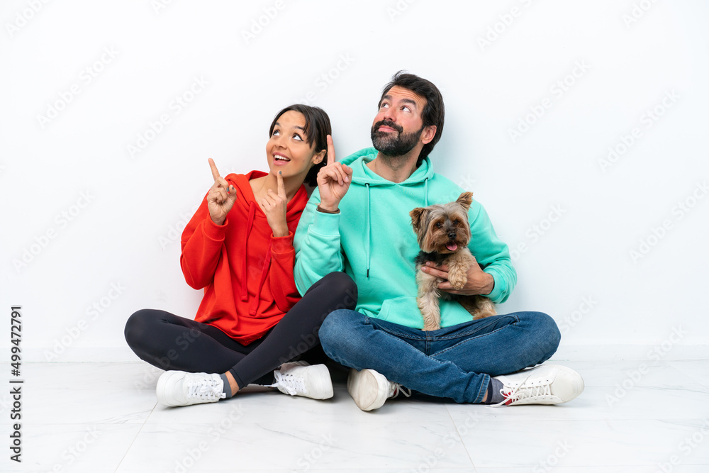 Young caucasian couple sitting on the floor with their pet isolated on white background pointing up with the index Young caucasian couple sitting on the floor with their pet isolated on white background pointing up with the index