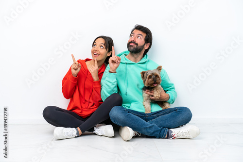 Young caucasian couple sitting on the floor with their pet isolated on white background pointing up with the index