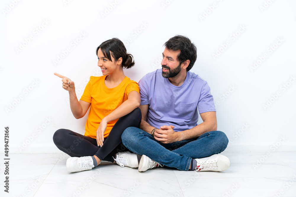 Young caucasian couple sitting on the floor isolated on white background pointing to the side to present a product