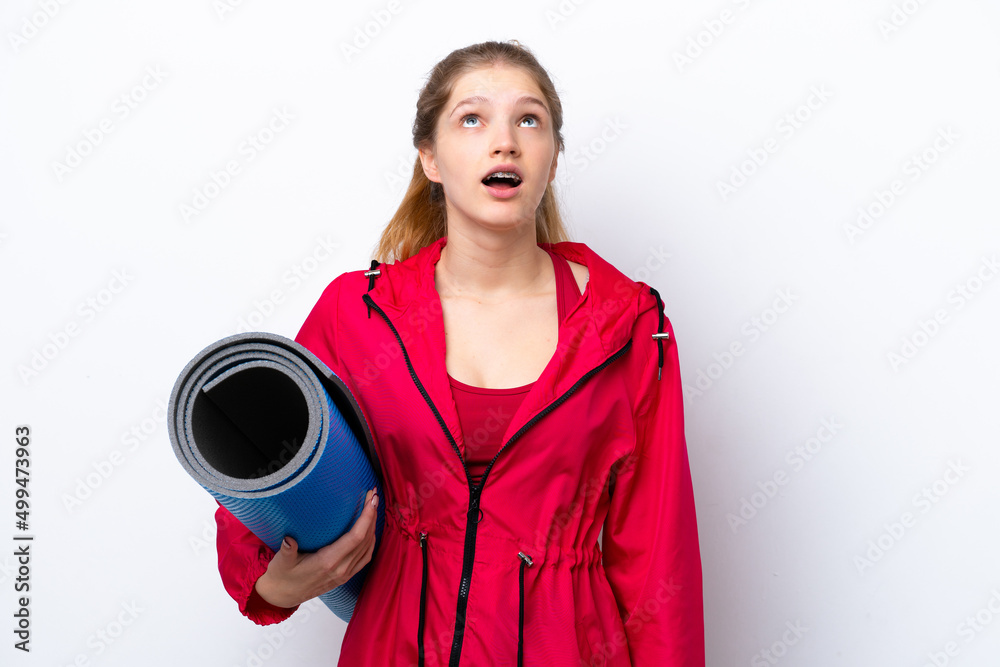 Teenager girl going to yoga classes while holding a mat isolated on white bakcground looking up and with surprised expression Teenager girl going to yoga classes while holding a mat isolated on white bakcground looking up and with surprised expression