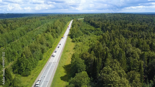 A wonderful green road from a helicopter . Clip. A long paved road with cars and a huge green summer forest around.