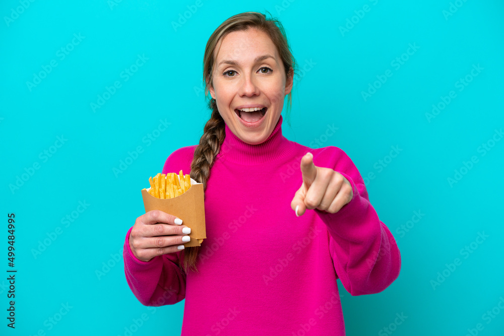 Young caucasian woman holding fried chips isolated on blue background surprised and pointing front Young caucasian woman holding fried chips isolated on blue background surprised and pointing front