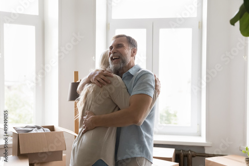 Loving older husband cuddles his hoary wife congratulates her move day end, mature family celebrate relocation standing near boxes with stuff, feel happy. Bank loan, new life, relations, love concept