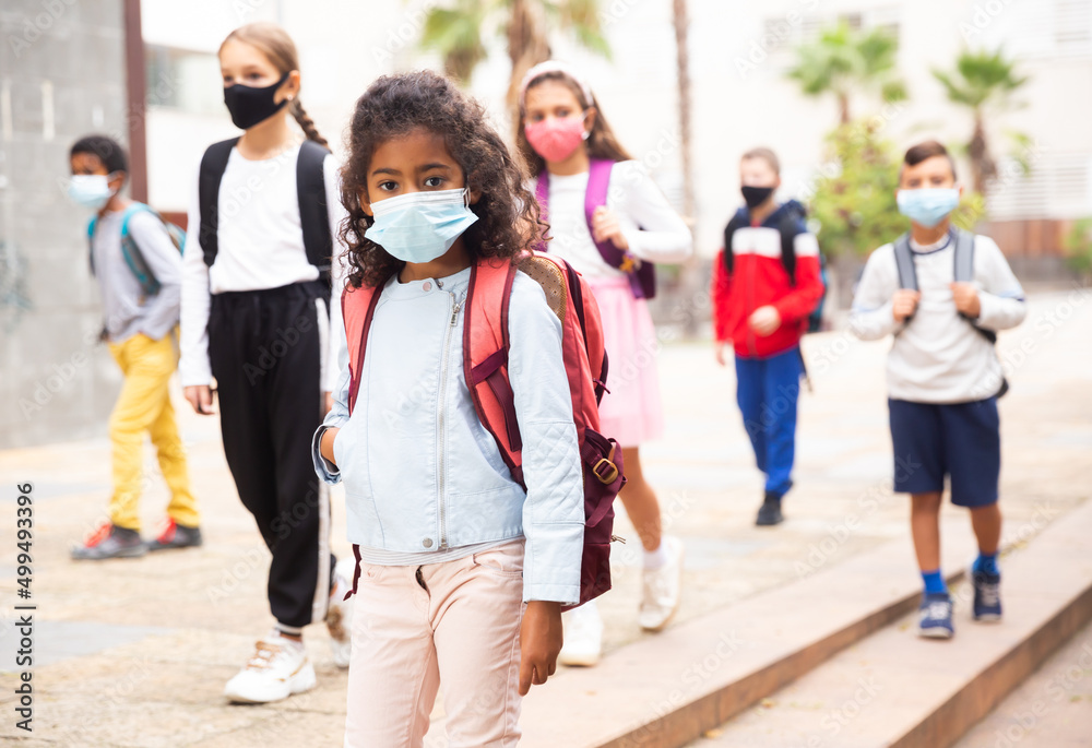 Obraz premium Portrait of positive schoolgirl in mask standing near school, children on background