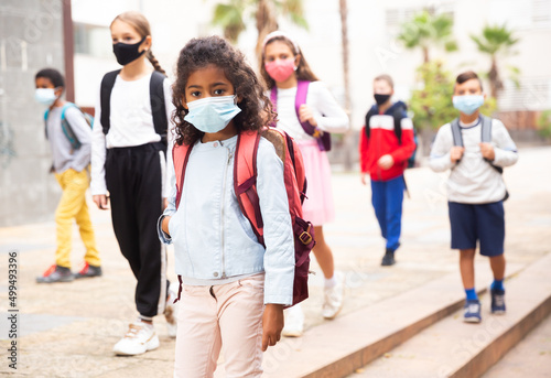 Portrait of positive schoolgirl in mask standing near school, children on background