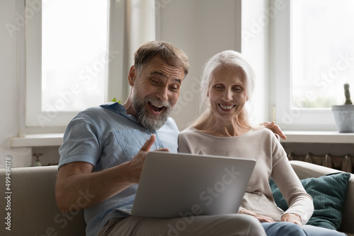 Smiling elderly couple watch movie funny videos look overjoyed, read fantastic news feels excited staring at laptop screen relax on sofa at home. Celebrate online lottery, giveaway win success concept