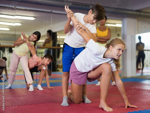 Young boys and girls training self-defence moves during group training. Female trainer standing nearby and observing.