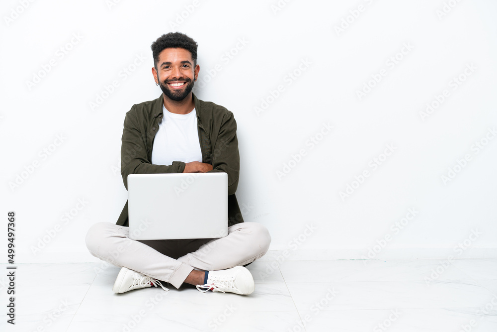Young Brazilian man with a laptop sitting on the floor isolated on white keeping the arms crossed in frontal position