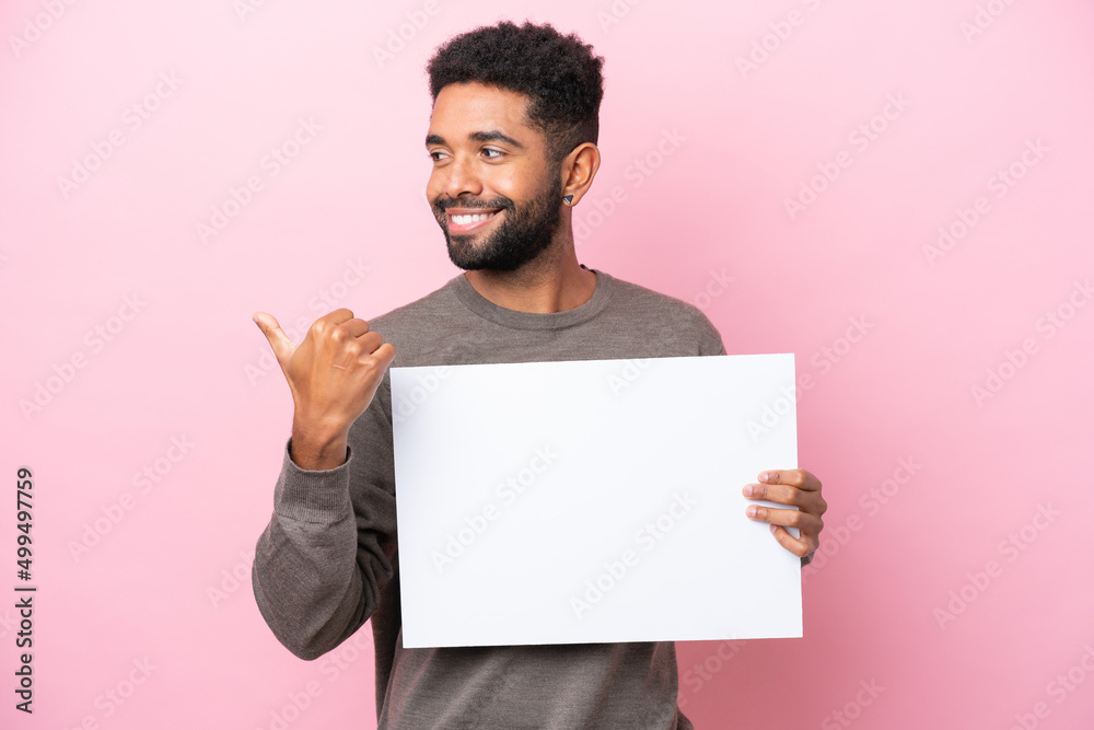 Young Brazilian man isolated on pink background holding an empty placard and pointing side Young Brazilian man isolated on pink background holding an empty placard and pointing side