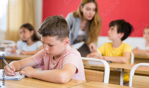 Portrait of concentrated tween boy studying in classroom, listening to schoolteacher and writing in notebook
