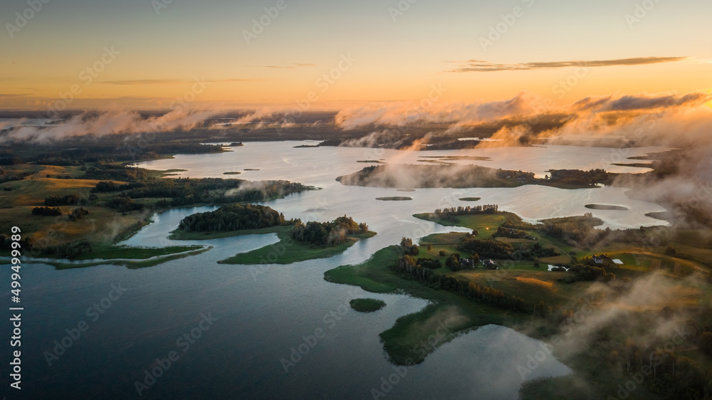 Lake land view from above, drone shot over beautiful islands and low clouds moving in wind backlit with morning sunlight. Aerial, flight over lakes during fantastic sunrise. Warm skyline on horizon