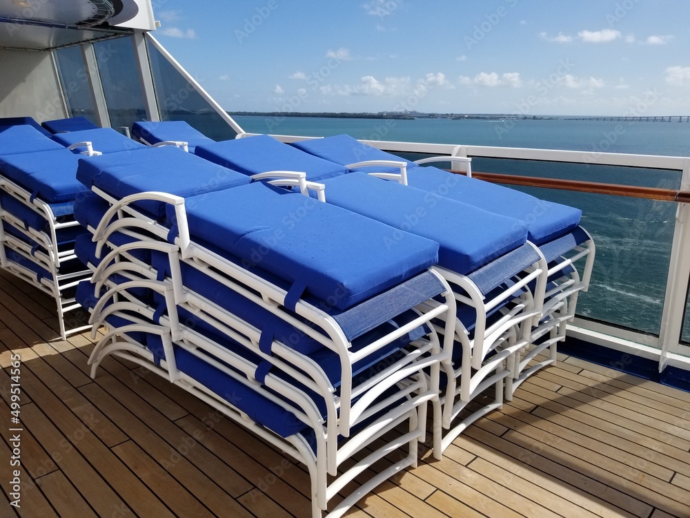 Lounge chairs with blue cushions folded up on cruise ship wooden deck with ocean in background 