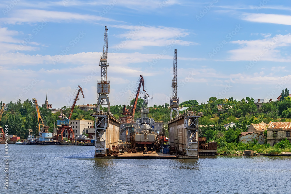 Repair platform of Mykolayiv Shipyard - view from the Pedestrian bridge across the Inhul river in Mykolaiv, Ukraine. Workshops and harbor cranes against the backdrop of a green shore on a summer day
