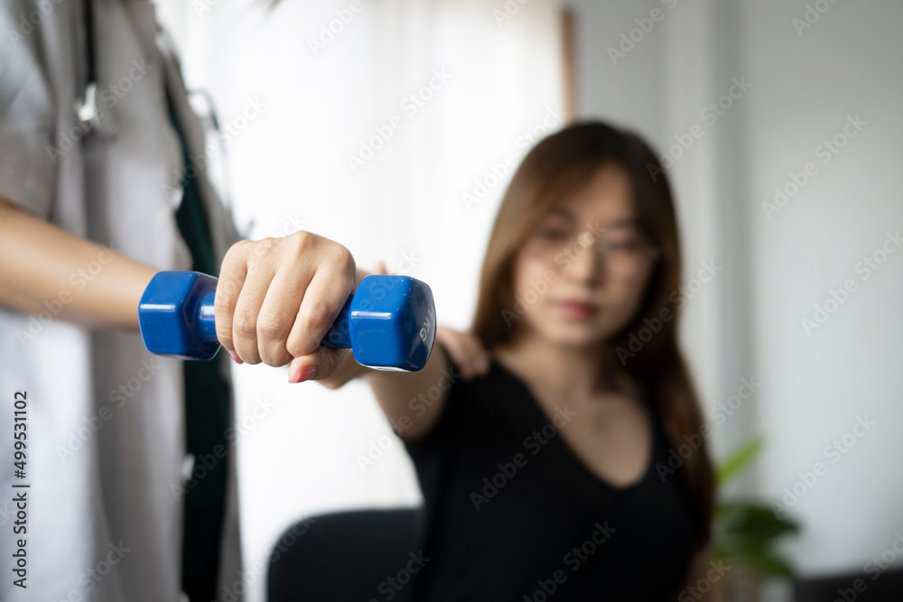 Fototapeta premium Young woman patient using dumbbells with outstretched arms while training with physiotherapist at clinic.