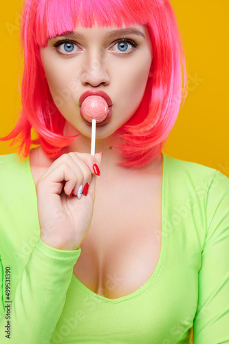 Hipster woman in pink wig with beautiful breasts eat lick lollipop on bright yellow background