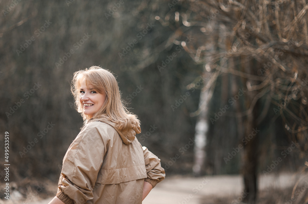 A beautiful happy middle-aged blonde woman in a beige jacket walks through a sunny spring or winter park. The woman looks around and laughs. Happiness and mental health. A beautiful happy middle-aged blonde woman in a beige jacket walks through a sunny spring or winter park. The woman looks around and laughs. Happiness and mental health.
