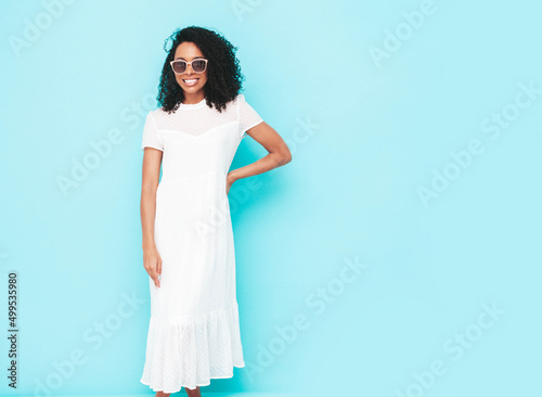Beautiful black woman with afro curls hairstyle. Smiling model dressed in white summer dress. Sexy carefree female posing near blue wall in studio. Tanned and cheerful in sunglasses. Isolated © halayalex