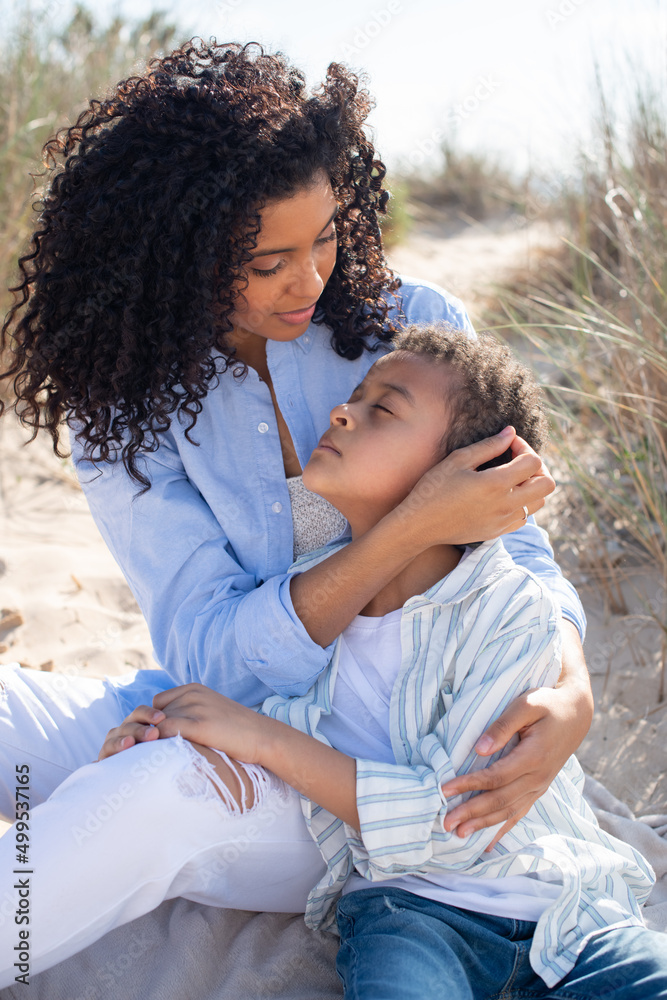 Fototapeta premium Caring African American mother and child on beach. Mother and son in casual clothes sitting on blanket, hugging, eyes closed. Family, relaxation, nature concept