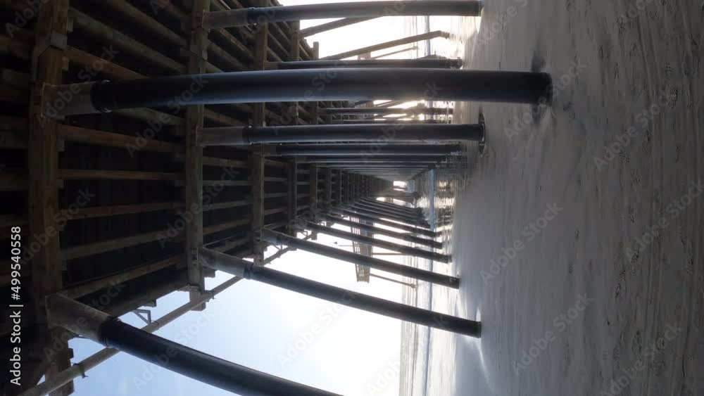 Walking under a wooden pier leading from the beach to the ocean - vertical orientation in slow motion Walking under a wooden pier leading from the beach to the ocean - vertical orientation in slow motion