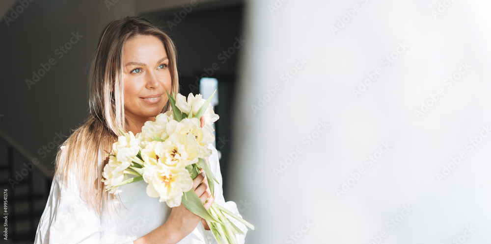 Young smiling woman with blonde long hair in white shirt with bouquet of yellow flowers in hands near window in bright interior, banner Young smiling woman with blonde long hair in white shirt with bouquet of yellow flowers in hands near window in bright interior, banner