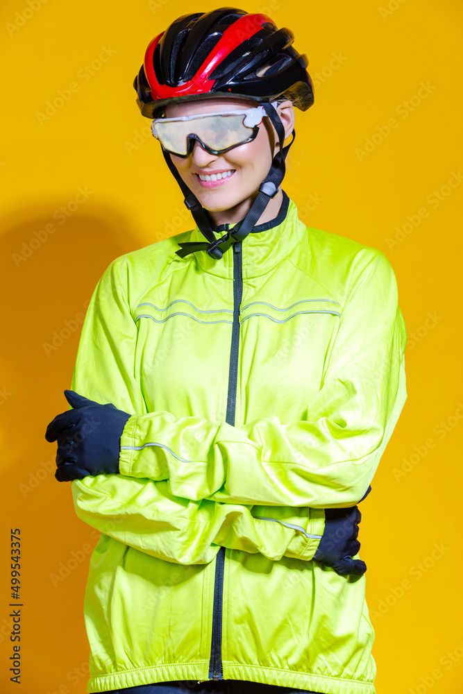 Fototapeta premium Women in Sport Concepts. Winsome Road Cyclist in Professional Warm Outfit Standing In Green Autumn Jersey and Facial Mask Goggles Against Yellow Background.