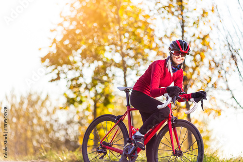 Fototapeta Naklejka Na Ścianę i Meble -  Professional Road Cycling. Winsome Caucasian Female Cyclist In Warm Outfit Posing On Road Bike While Getting Ready for Start Outdoors Against Autumn Background