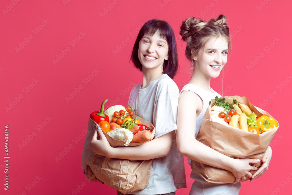Couple of Smiling Laughing Positive Caucasian Girlfriends With Disposable paper Eco Bags Filled With Grocery And Vegetables Over Pink Rose Background. Couple of Smiling Laughing Positive Caucasian Girlfriends With Disposable paper Eco Bags Filled With Grocery And Vegetables Over Pink Rose Background.