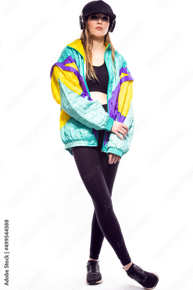 Full Length Portrait of Winsome Sportwoman in Fitness Outfit and Baseball Cap With Wireless Headphones Listening Music While Looking Straight With Hands Crossed In Front Over White. Full Length Portrait of Winsome Sportwoman in Fitness Outfit and Baseball Cap With Wireless Headphones Listening Music While Looking Straight With Hands Crossed In Front Over White.