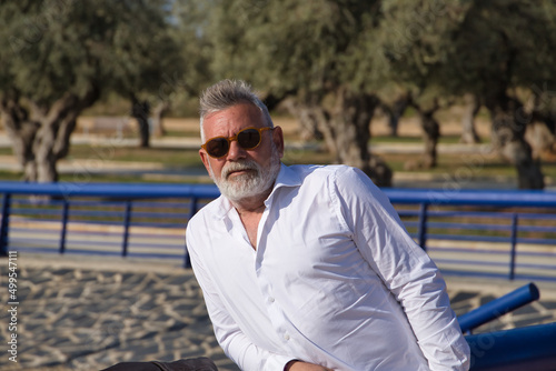 Mature man, gray-haired, bearded, sunglasses, white shirt, leaning on a railing outdoors. Concept grandfather, sugar daddy, lifestyle, mature man.
