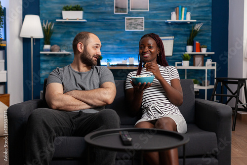 Happy interracial couple having a pleasent conversation while sitting on sofa at home. Relaxed man and woman bonding and having a good time in the living room while sharing snacks photo