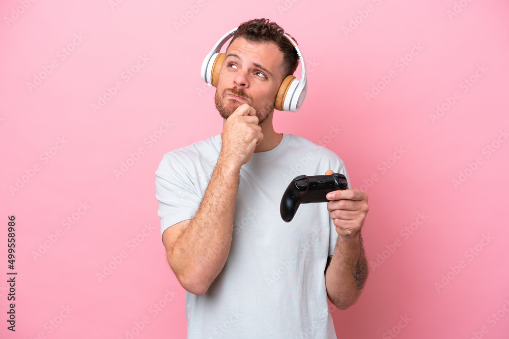Naklejka premium Young Brazilian man playing with video game controller isolated on pink background having doubts and thinking