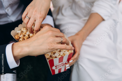 Wedding. The groom in a suit and white shirt and the bride in a white dress are sitting and eating popcorn from red boxes