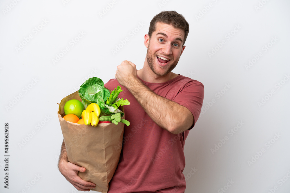 Young caucasian man holding a grocery shopping bag isolated on white background celebrating a victory Young caucasian man holding a grocery shopping bag isolated on white background celebrating a victory