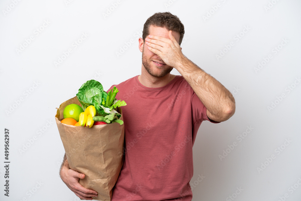 Young caucasian man holding a grocery shopping bag isolated on white background covering eyes by hands. Do not want to see something Young caucasian man holding a grocery shopping bag isolated on white background covering eyes by hands. Do not want to see something
